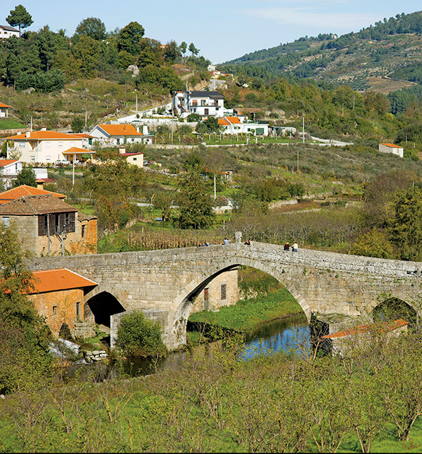 Lush green mountains and sprawling vineyards surrounding an ancient bridge in the village of Ucanha, Portugal.